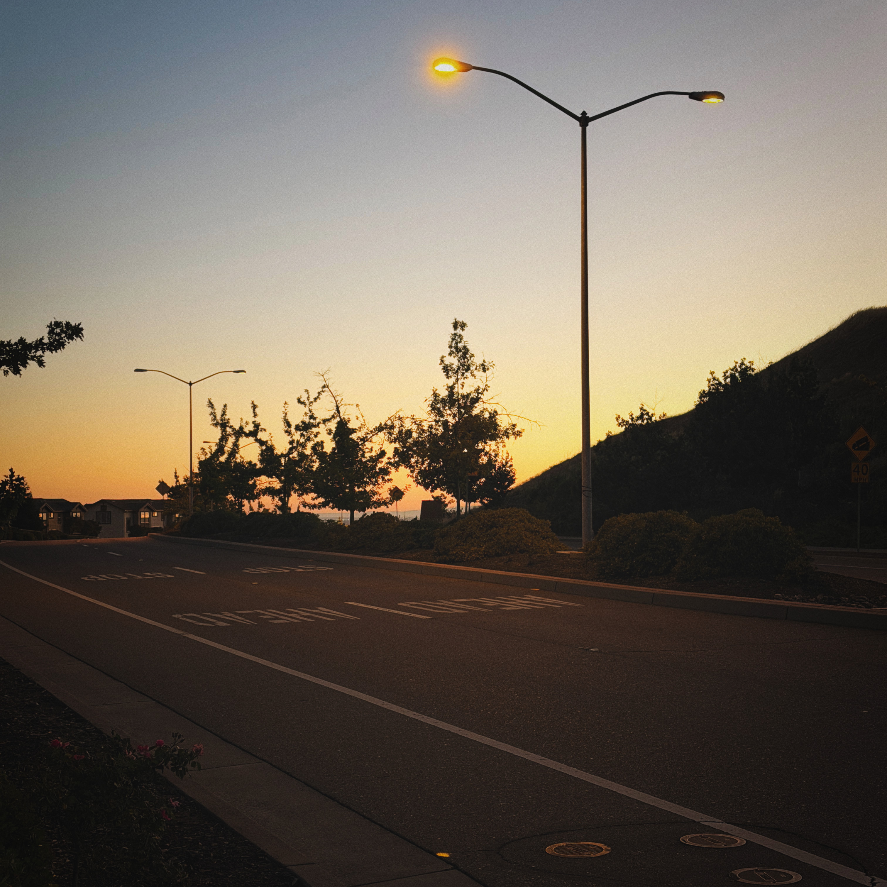 A quiet road at sunset with glowing streetlights, trees, a hillside, and warm light near the horizon.
