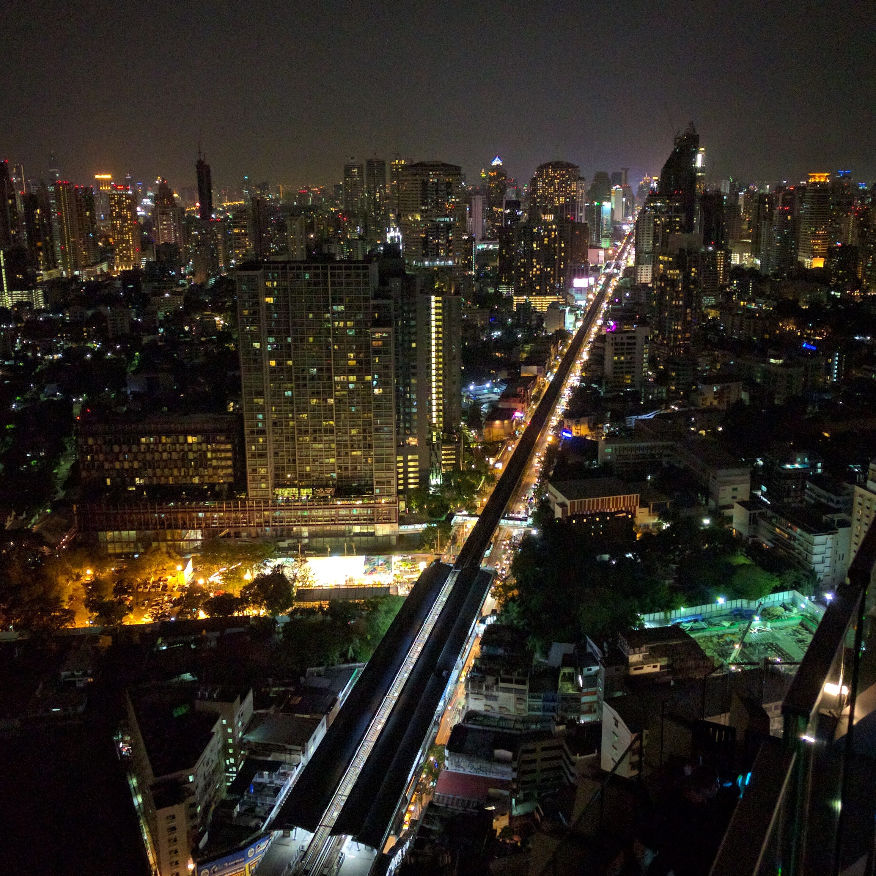 A nighttime view over Bangkok with lit high-rise buildings, traffic, and an elevated train line cutting through the city.