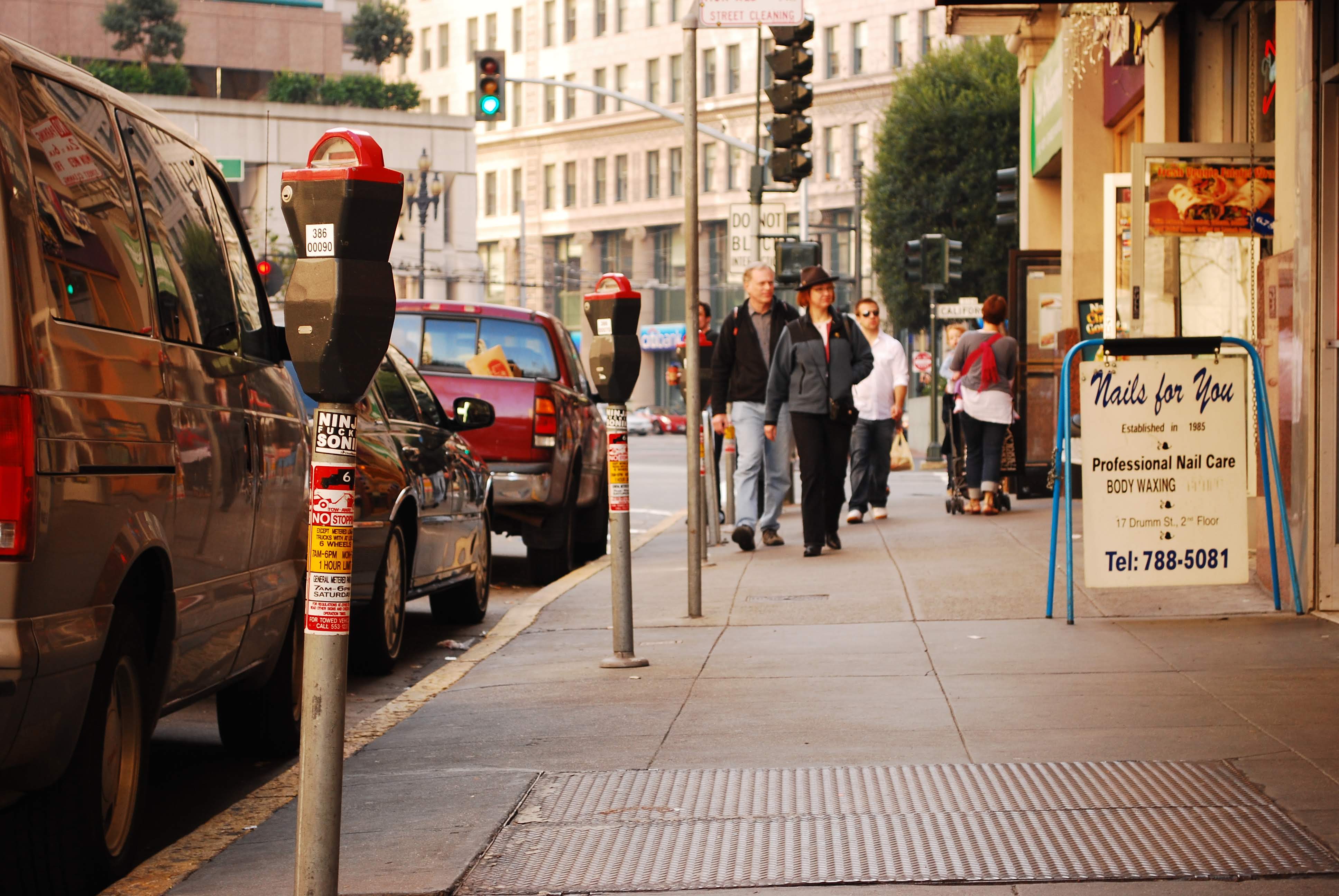 A city sidewalk scene with parking meters, parked cars, pedestrians, and a storefront sign for a nail salon.