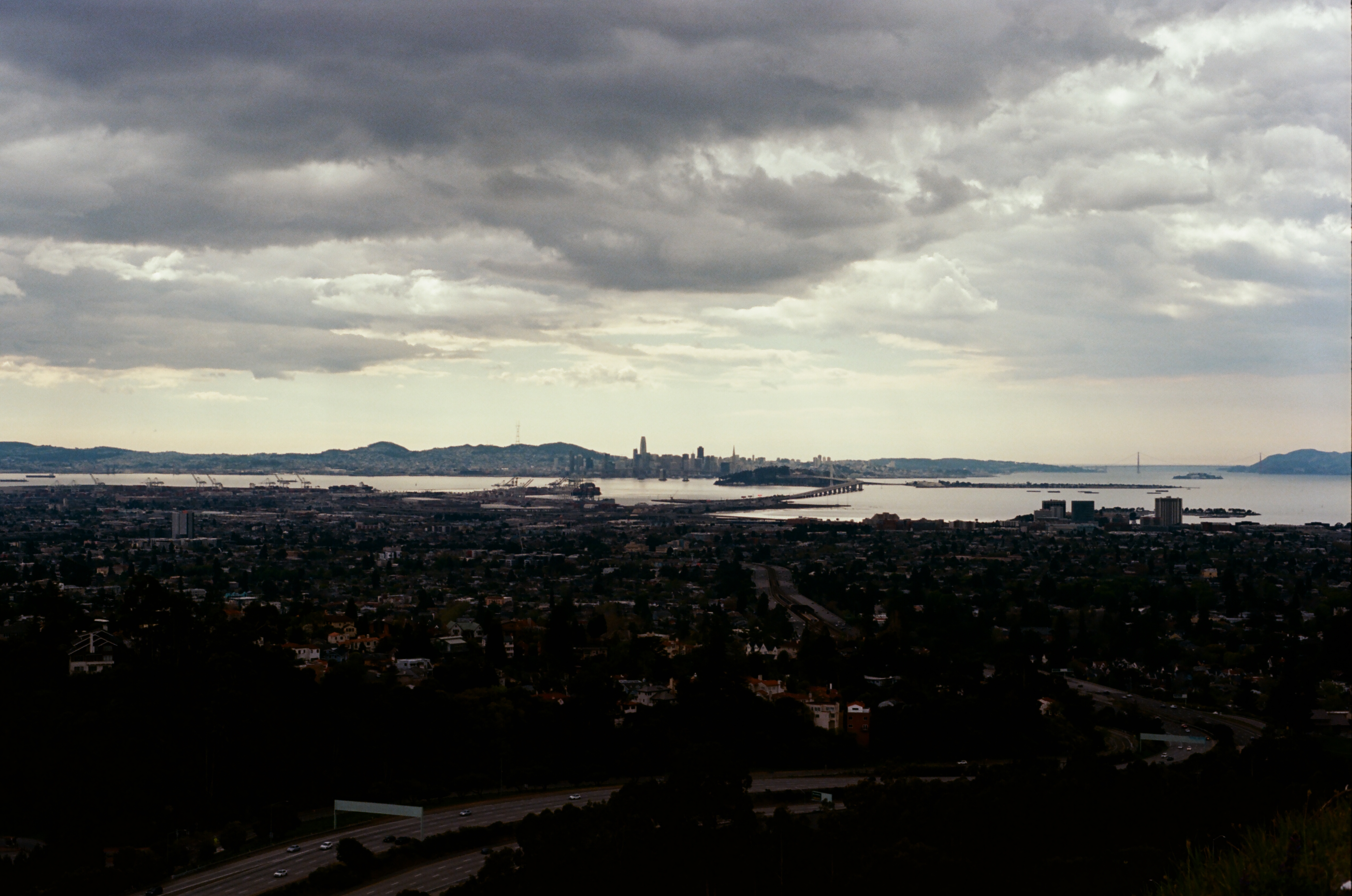 A wide film photograph of the San Francisco Bay Area under heavy clouds, with the city skyline and Bay Bridge in the distance.